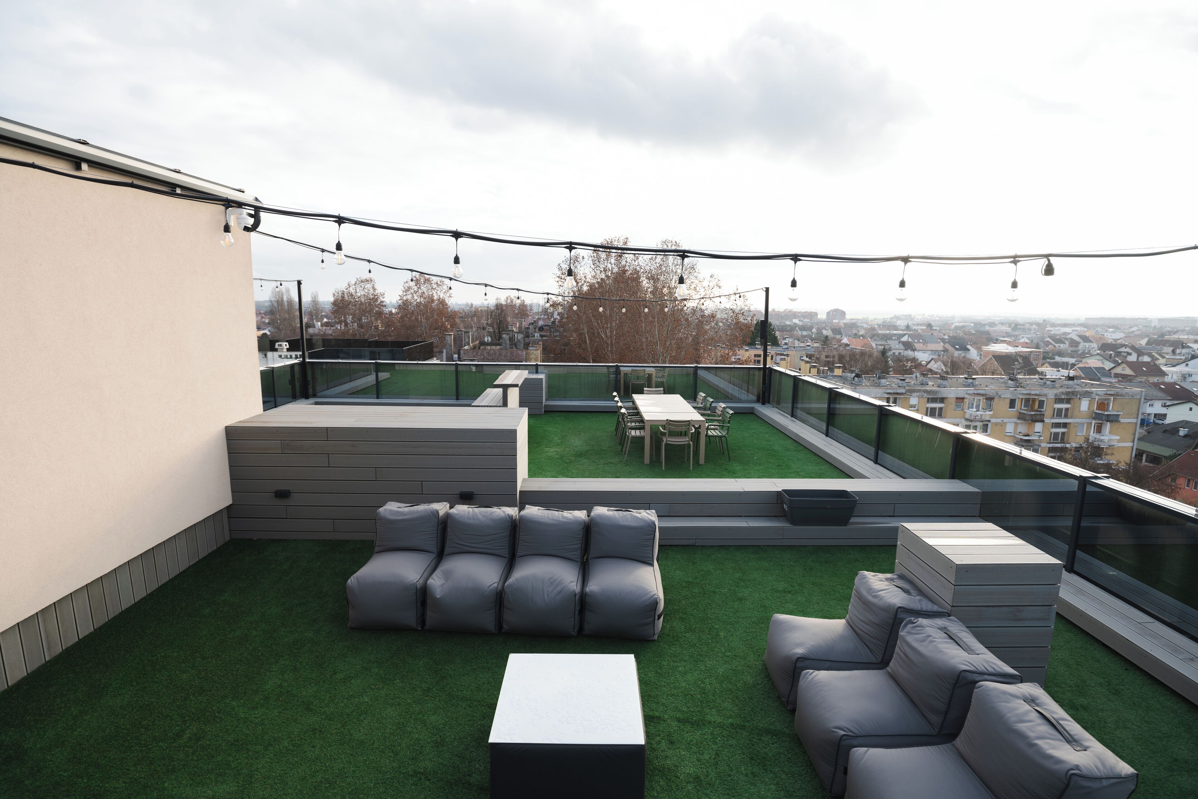 A rooftop terrace with artificial grass, grey sofas, a dining table, and string lights.