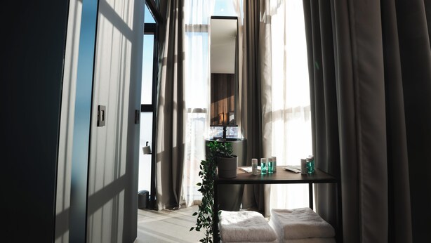 A modern hotel room with a large mirror, a bedside table with toiletries, and a view of the city through large windows. - DISTRICT boutique  (Osijek)