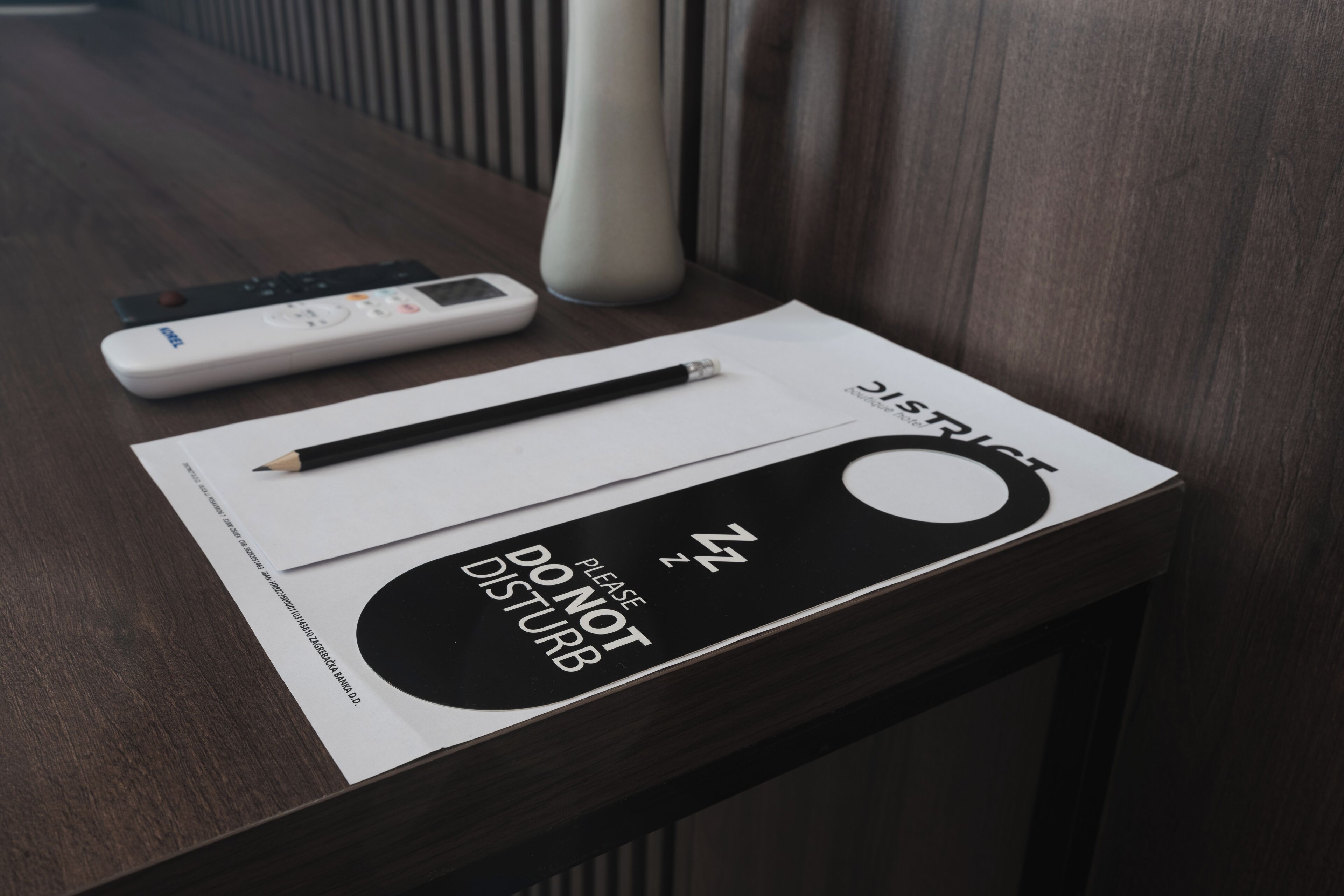 A wooden table with a remote control, a pencil, and a booklet titled "DISTRICT PLEASE DO NOT DISTURB".