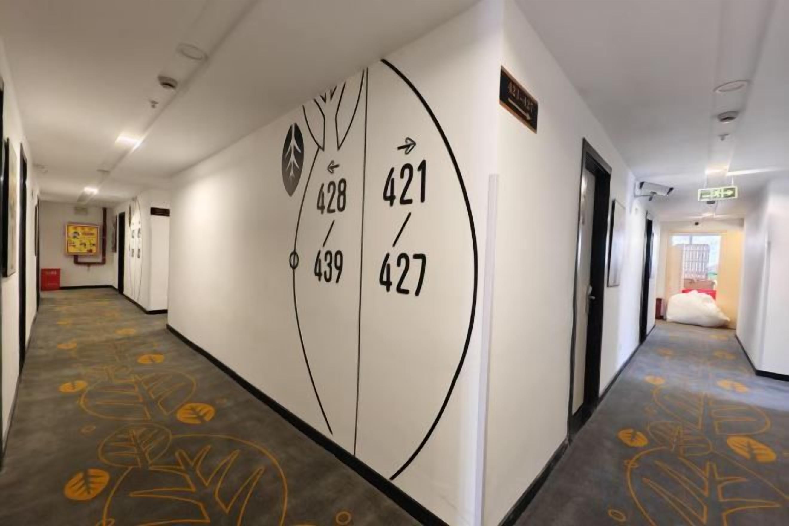 Un pasillo de hotel con alfombra de motivos vegetales, una pared con números de habitaciones y una puerta a la derecha.