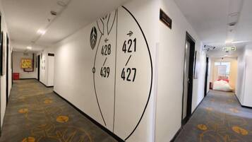 Un pasillo de hotel con alfombra de motivos vegetales, una pared con números de habitaciones y una puerta a la derecha.
