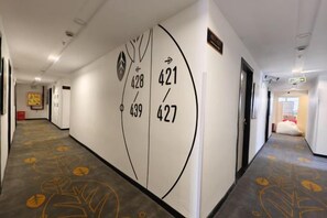 Corredor de hotel com carpete estampado com motivos de folhas, parede com números de quarto e uma porta à direita.