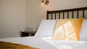 Een net opgemaakt bed met witte en gele kussens, een houten hoofdbord en een nachtkastje met een lamp.