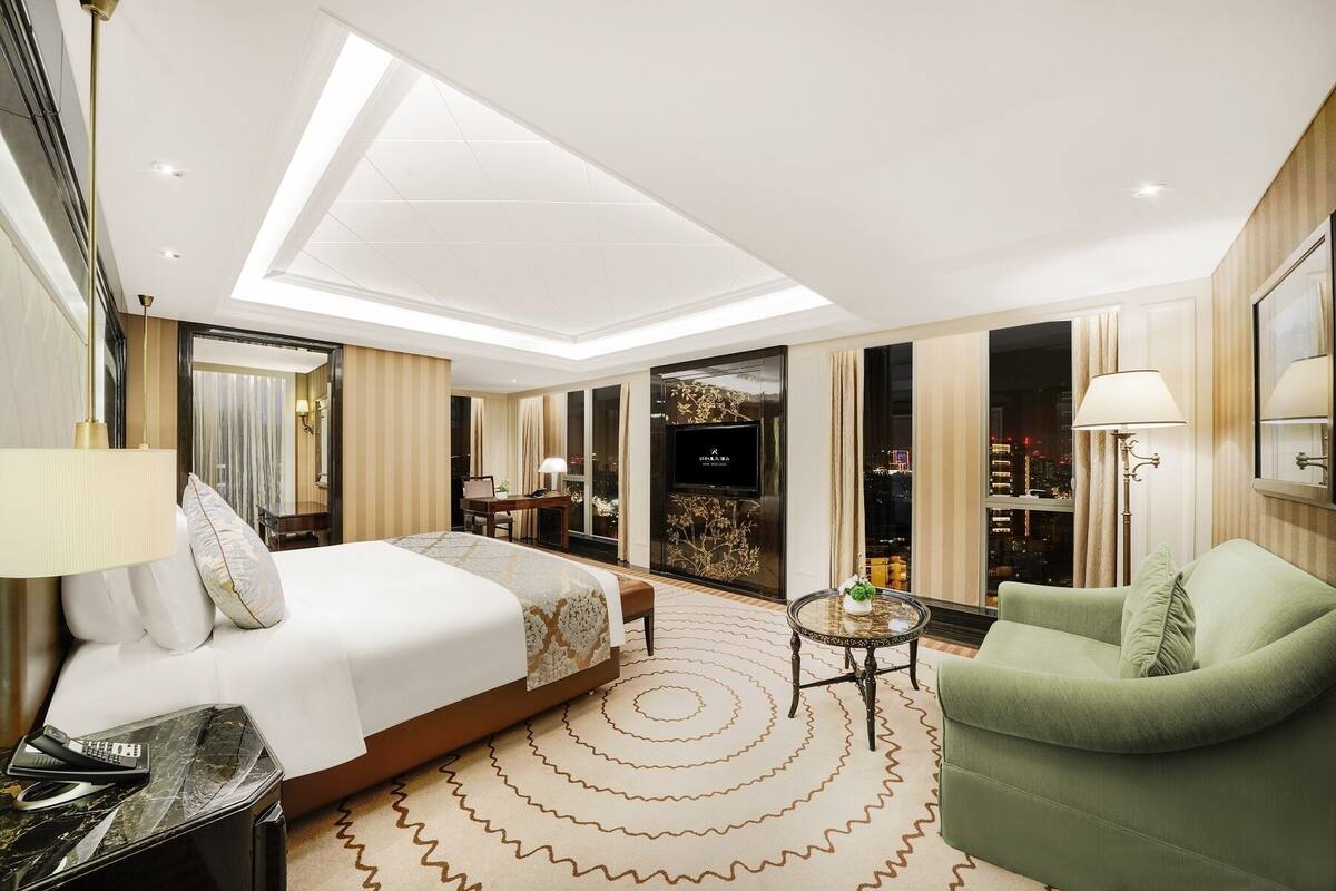Quarto de hotel moderno com uma cama grande, uma poltrona verde, uma mesa pequena e uma televisão na parede.