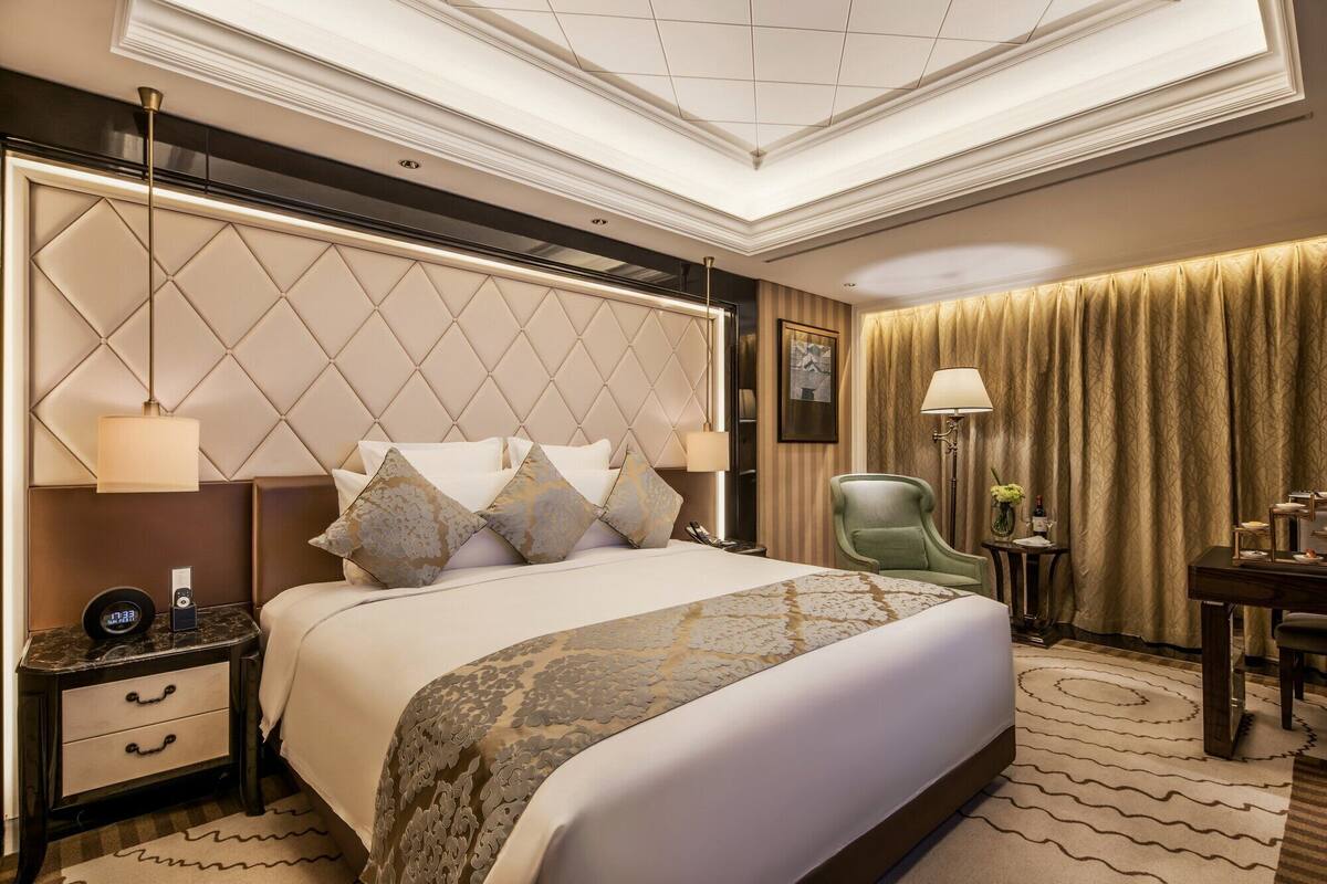 Quarto de hotel com uma cama grande, uma mesinha de cabeceira com um relógio, uma poltrona verde, uma mesa lateral com abajur e um tapete estampado.