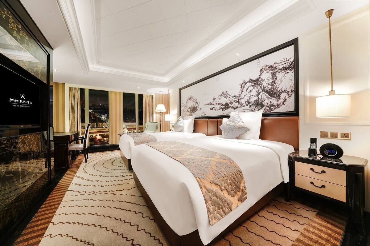 Quarto de hotel com uma cama grande, mesa de cabeceira, televisão e vista para a cidade à noite.
