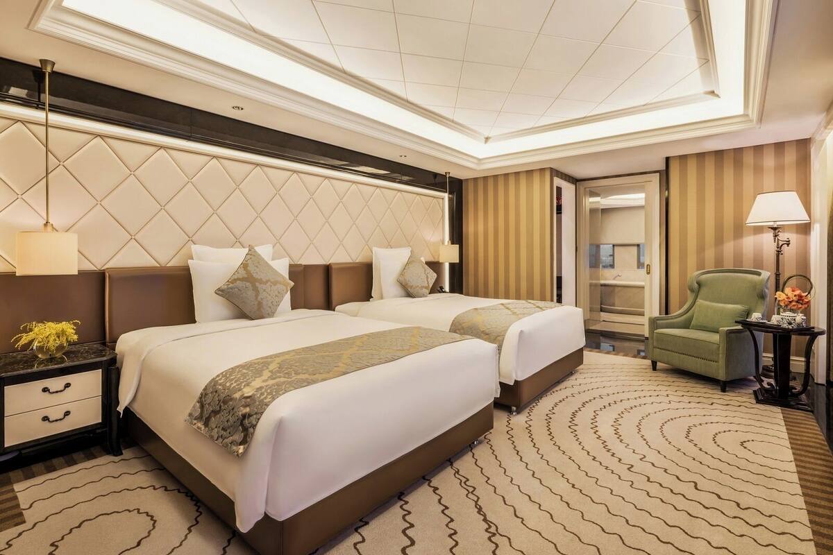 Quarto de hotel com duas camas, uma poltrona verde, uma mesinha com luminária e uma janela grande.