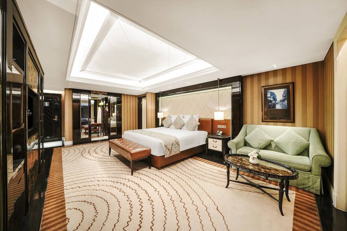 Quarto de hotel com uma cama grande, uma poltrona verde, uma mesinha e um espelho.