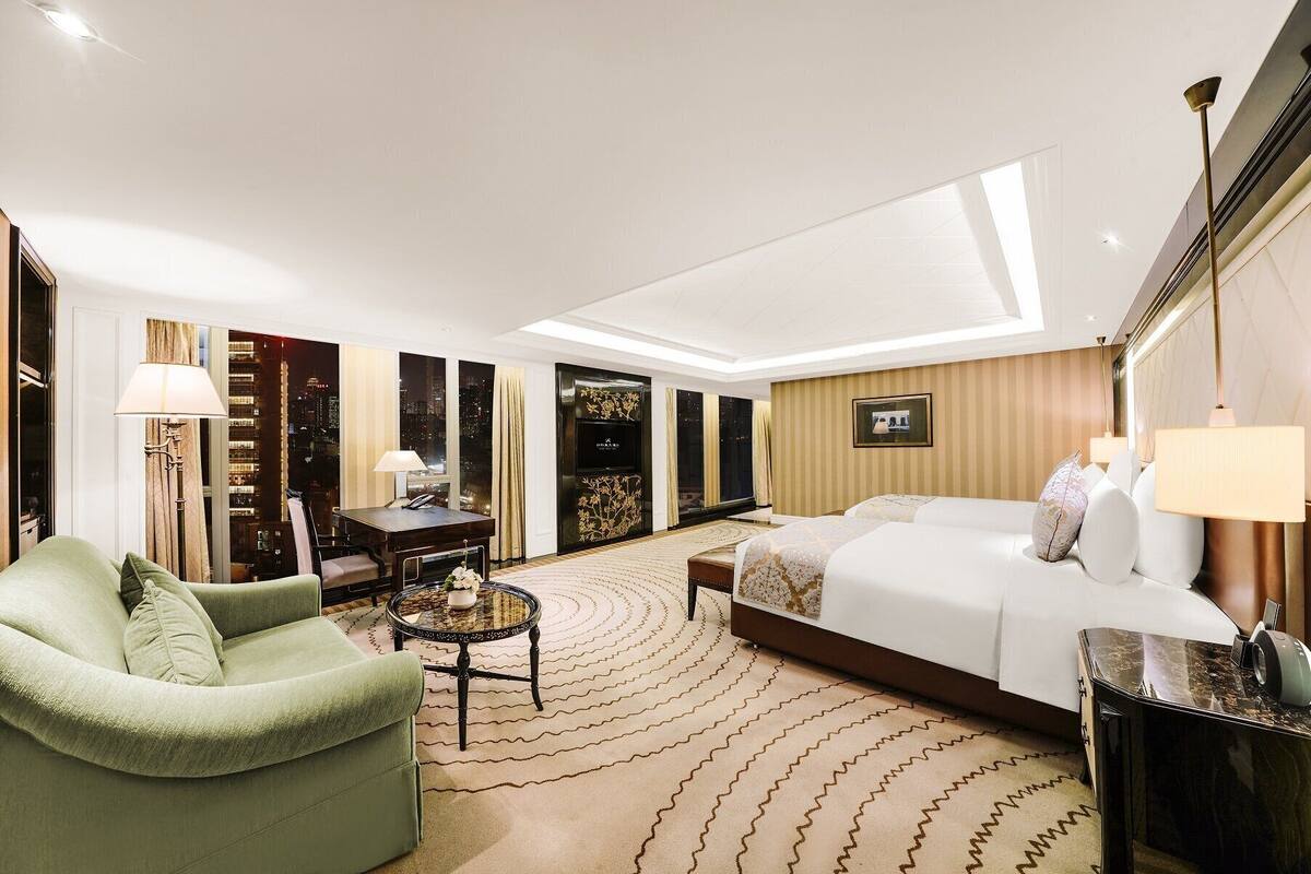 Quarto de hotel com cama, poltrona verde, escrivaninha com abajur e vista para a cidade à noite.