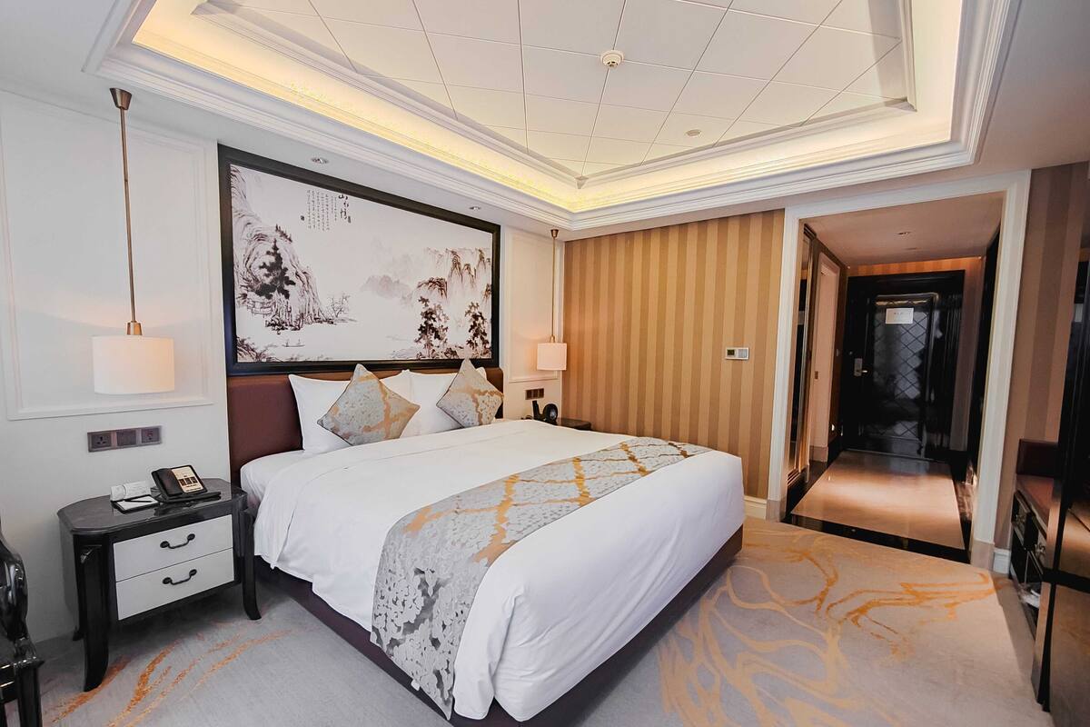 Quarto de hotel com uma cama grande, uma mesa de cabeceira, um telefone e uma obra de arte na parede.