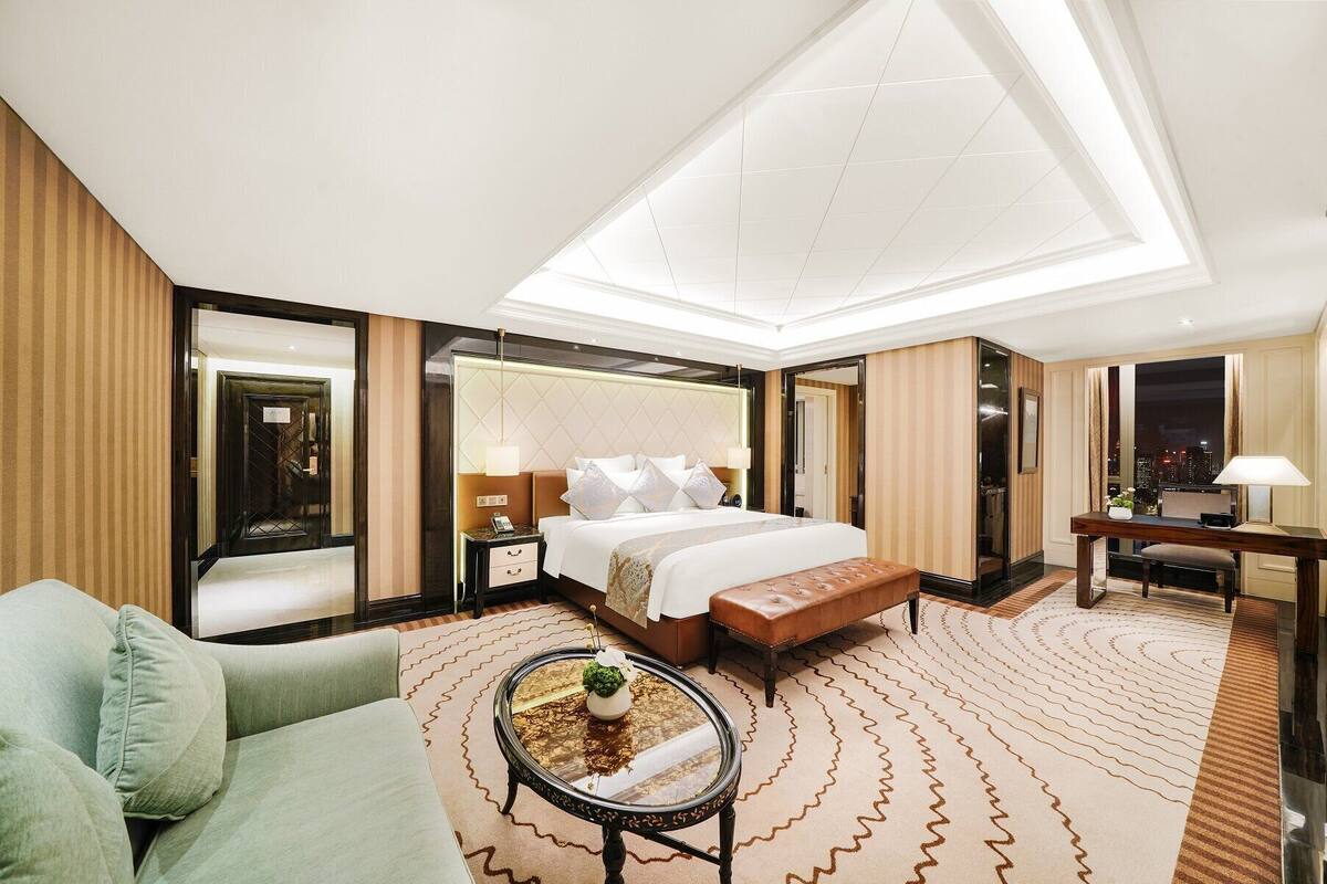 Quarto de hotel com uma cama grande, área de estar com sofá e mesa de centro, escrivaninha e vista para a cidade à noite.