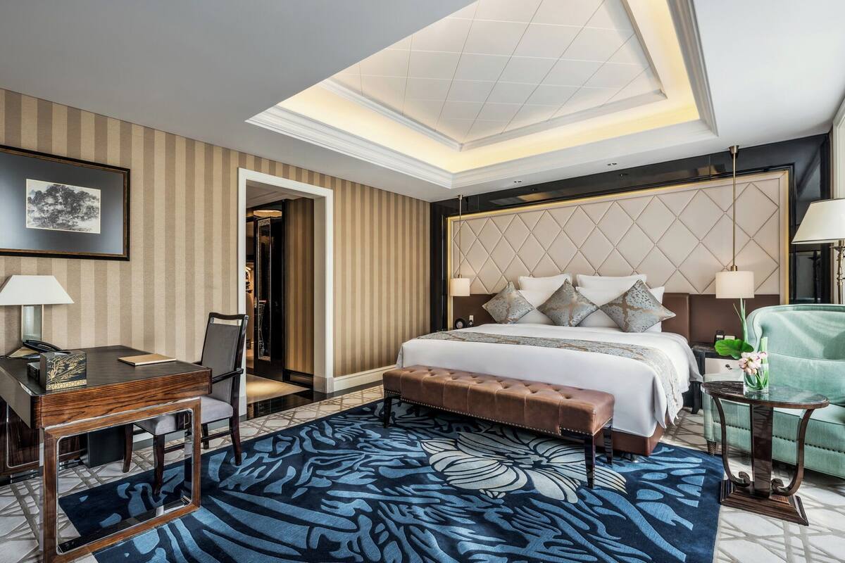 Quarto de hotel com uma cama grande, uma escrivaninha, uma poltrona verde e um tapete estampado.