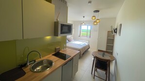 A compact kitchen with a microwave, sink, and wooden countertop. A bed with a window is visible in the background.