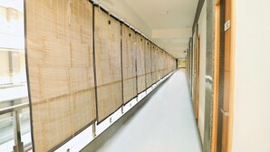 A long corridor with a series of rooms on one side, each with a wooden door and a window covered by a bamboo blind. - Hotel Santeria Inn (Surat)