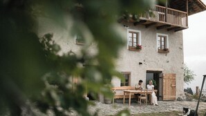 A rustic stone building with a wooden balcony, a wooden table and chairs outside, and a dog nearby.