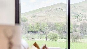 A room with a large window offering a view of a grassy hill and trees. - Balbeg Cottage by Interhome (Ayr)