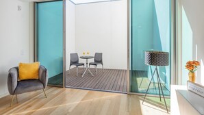 Una habitación moderna con una puerta de vidrio corredera que da a un balcón, una mesita con dos sillas y una lámpara de pie.