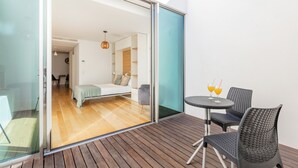 Habitación moderna con puerta de cristal que da a un balcón con suelo de madera, una cama y una mesita con dos bebidas.