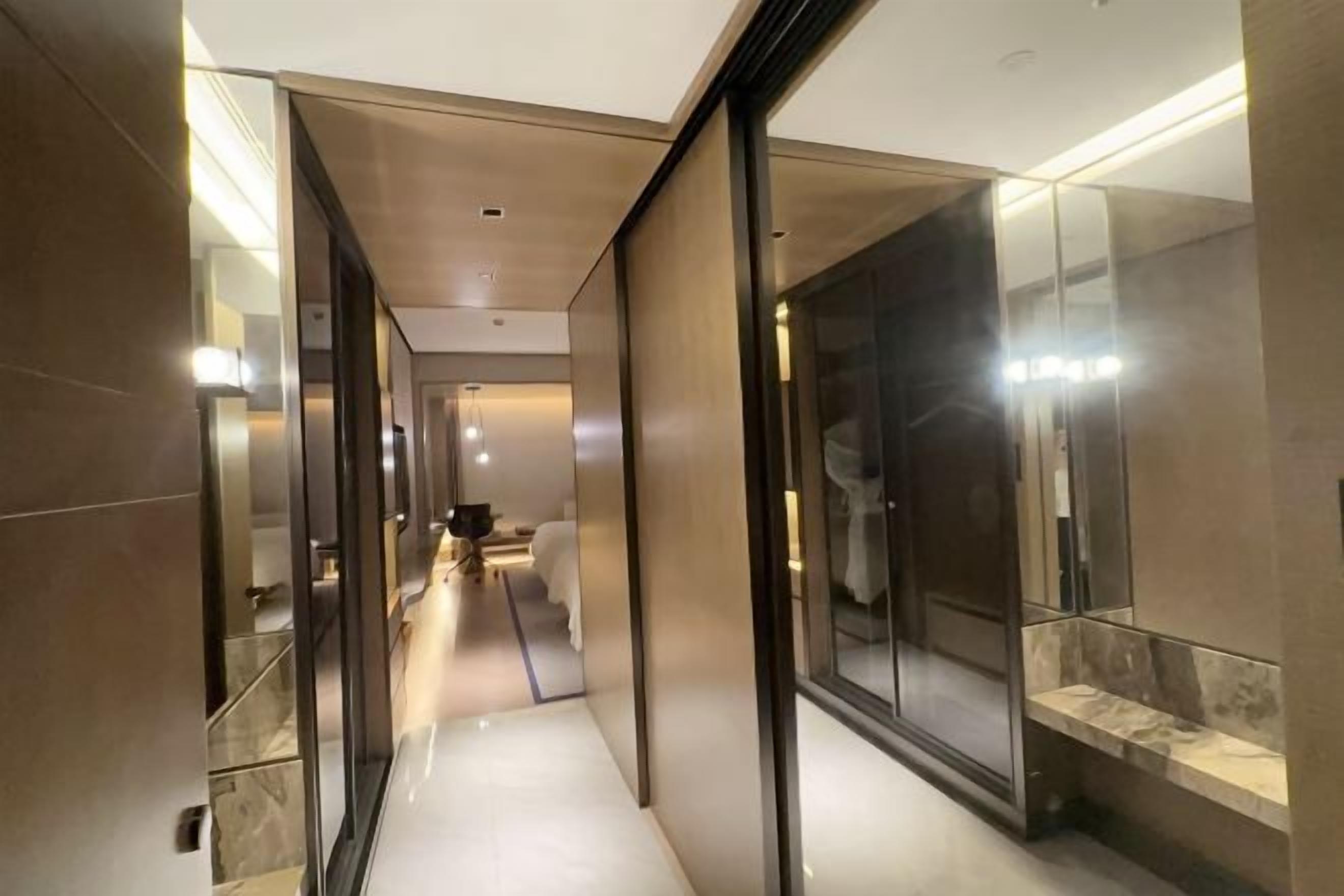 A modern hotel corridor with mirrored walls, a clean floor, and a well-lit hallway leading to a room with a bed and a desk.