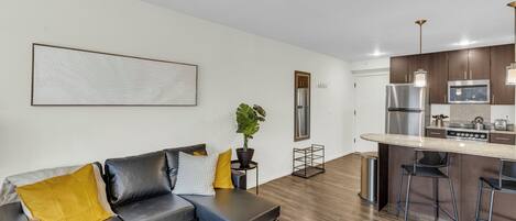 Una sala moderna con un sofá seccional gris, una mesita de centro pequeña y una planta en maceta. Se observa una cocina con una barra y taburetes al fondo.