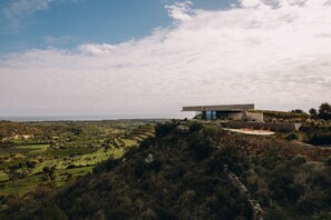 A modern house with a flat roof and large windows, surrounded by a hilly landscape with terraced fields.