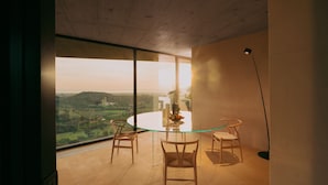 A modern dining area with a glass table and chairs, large windows offering a view of a hilly landscape, and a tall floor lamp. - Villa Esovisia by Elite Villas (Avola)