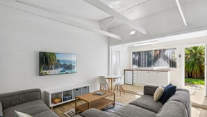 Una sala de estar moderna con un televisor de pantalla plana grande, sofás grises, una mesa de centro y una zona de cocina con ventana que da a un jardín.