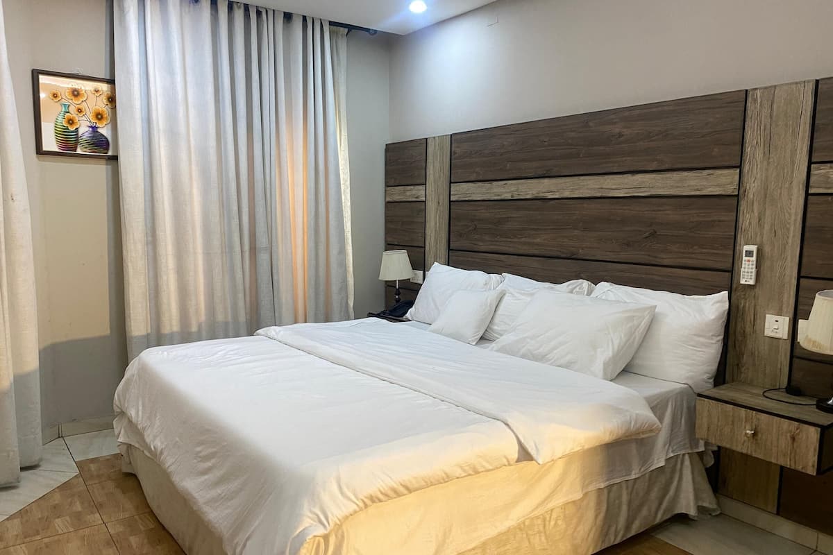 Un lit bien fait, avec du linge de lit blanc, une tête de lit en bois, une table de chevet avec une lampe et un téléphone fixé au mur.