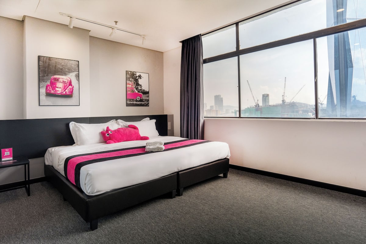 A hotel room with a large bed, a nightstand, two wall-mounted artworks, and a view of a cityscape through a large window.