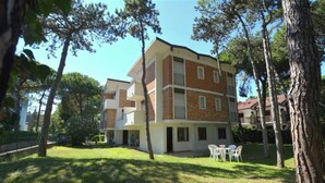 A multi-story building with a flat roof, surrounded by trees and a grassy area with outdoor seating. - Cozy Apartment With Fenced Garden in Lignano Sabbiadoro (Lignano Sabbiadoro)