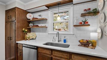 Een keuken met houten kastjes, een roestvrijstalen spoelbak en witte aanrechtbladen. Er zijn planken met decoratieve objecten en een raam boven de spoelbak.