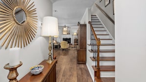 A hallway with a wooden floor, a white wall, a sunburst mirror, a lamp, a bowl, and a staircase.