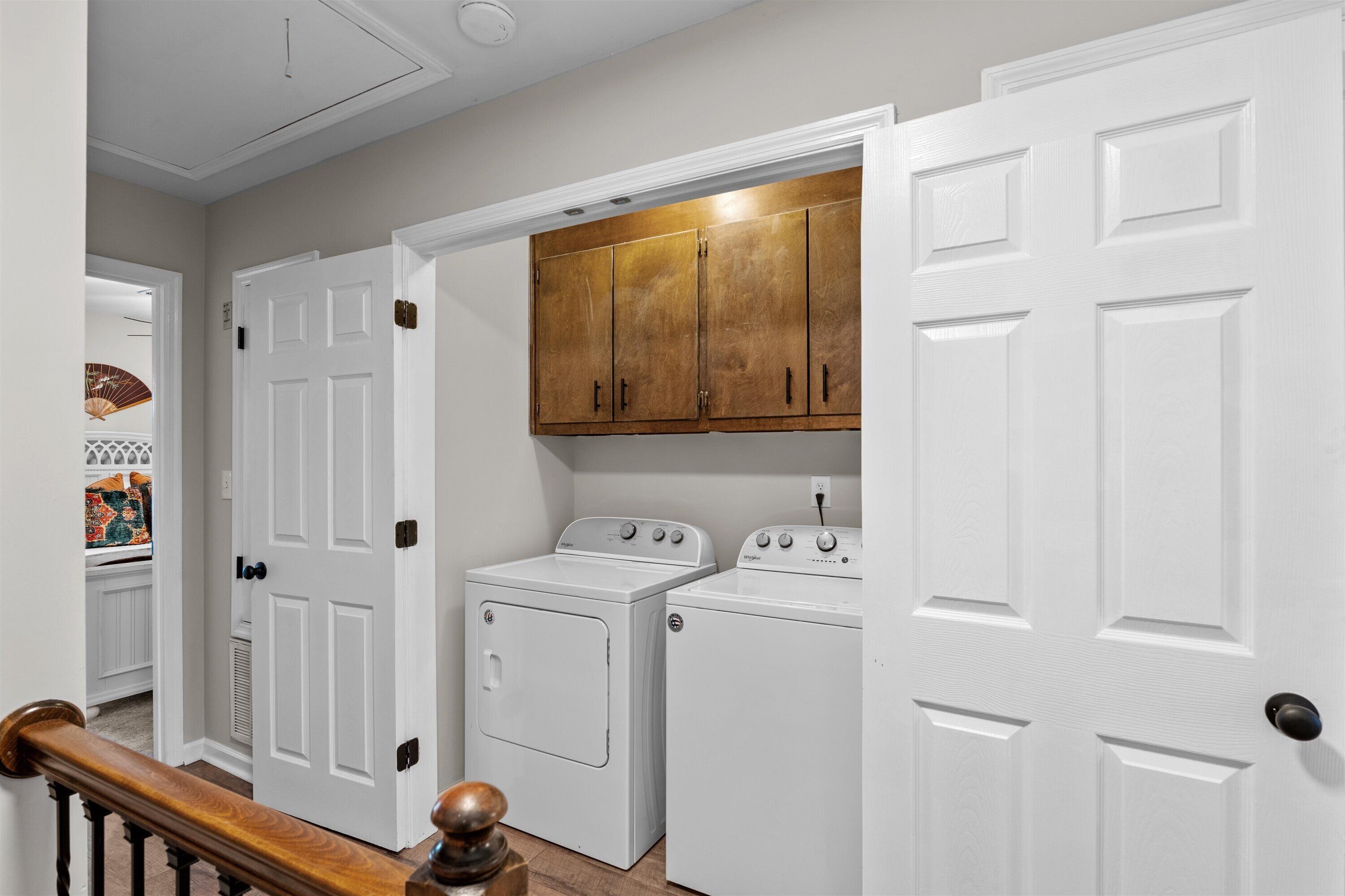 A laundry room with two white washers and dryers, wooden cabinets above, and a white door with a wooden handrail.