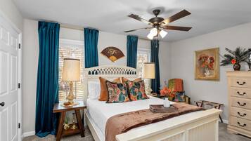 A bedroom with a bed, bedside tables, a ceiling fan, a window with curtains, a wall-mounted fan, and a dresser.