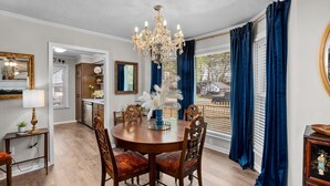 A dining area with a chandelier, wooden chairs, a round table, and a view through large windows with blue curtains.