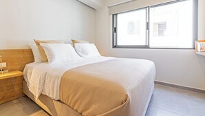A modern bedroom with a large bed, a wooden headboard, a bedside table, and a window with blinds. - Stone Haven Apt 2, By IdealStay Experience (Limenas Chersonisou, Greece)