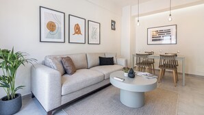 A modern living room with a grey sofa, a round coffee table, and a dining area with a table and chairs. There are framed artworks on the wall and a potted plant on the floor. - Stone Haven Apt 2, By IdealStay Experience (Limenas Chersonisou, Greece)