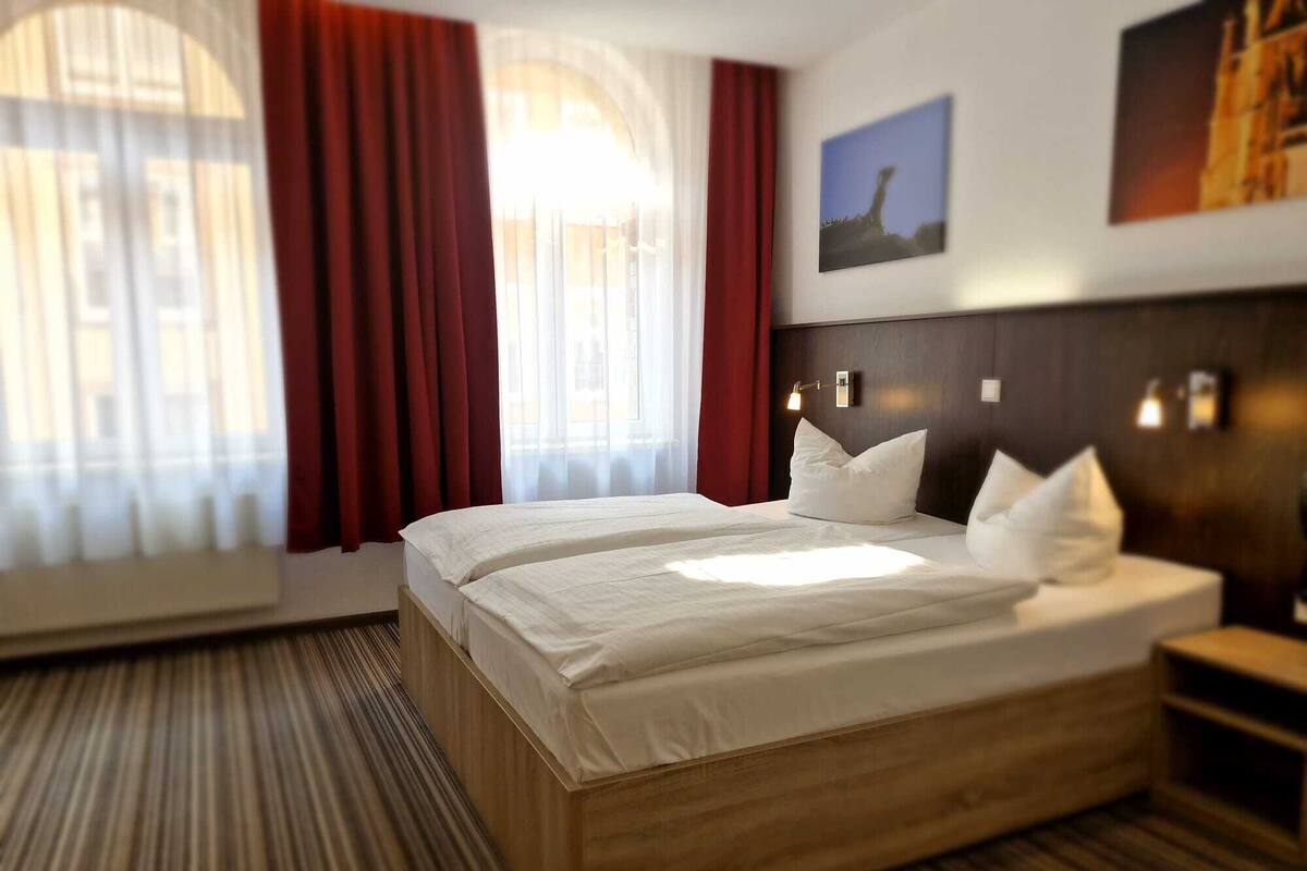 A hotel room with a large bed, two bedside tables, a nightstand, a window with curtains, and a wall-mounted light fixture.