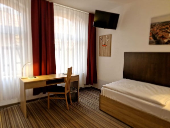 A hotel room with a bed, a desk, a chair, a TV mounted on the wall, and a window with curtains.
