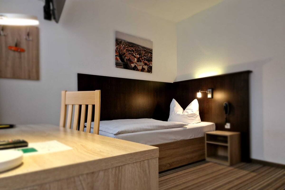 A hotel room with a wooden desk, a bed with white bedding, a dark headboard, a small bedside table, and a wall-mounted light fixture.