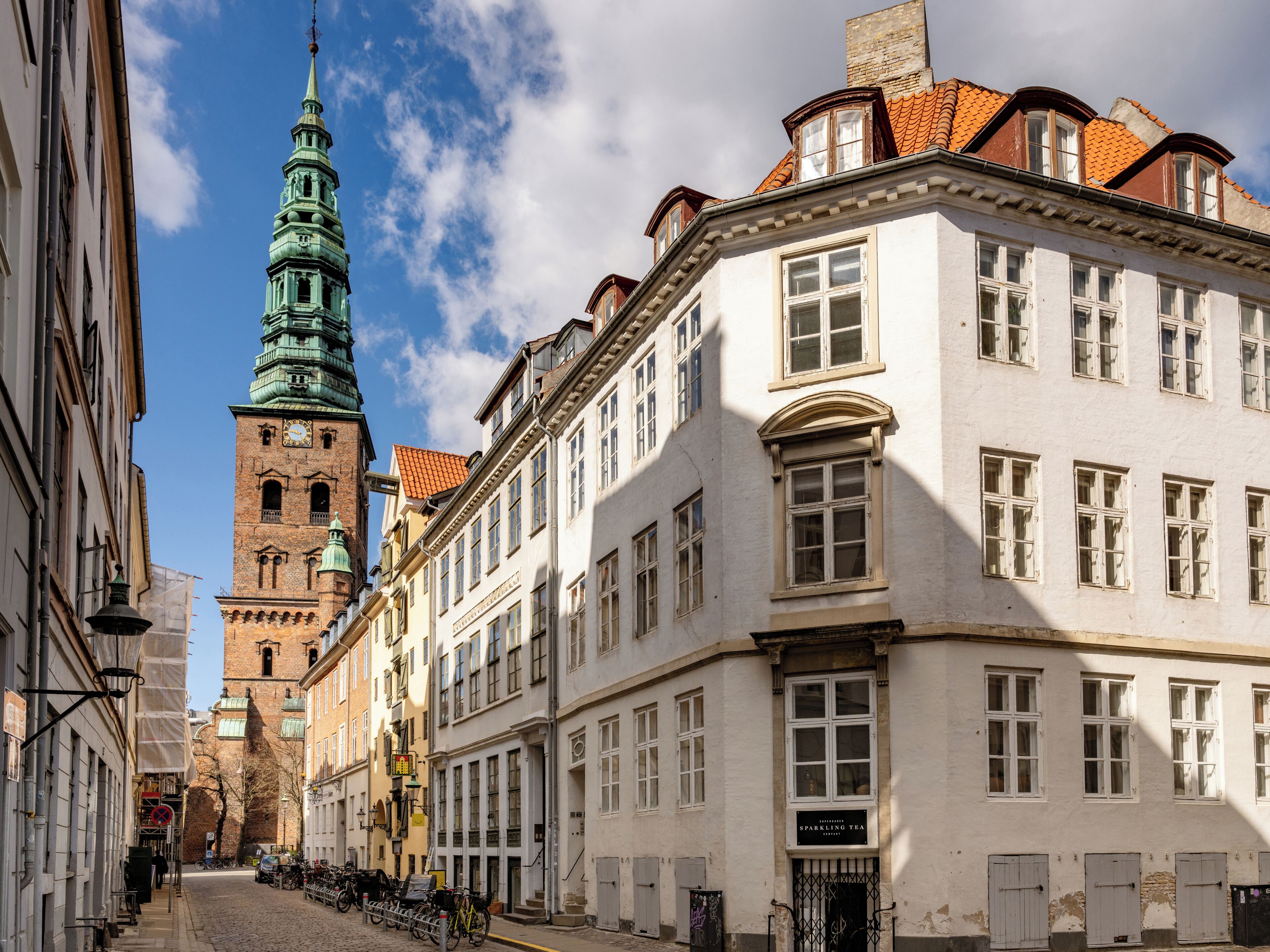 Vista de una calle con edificios históricos, una torre de iglesia y un cartel que dice 'Sparkling Tea'.