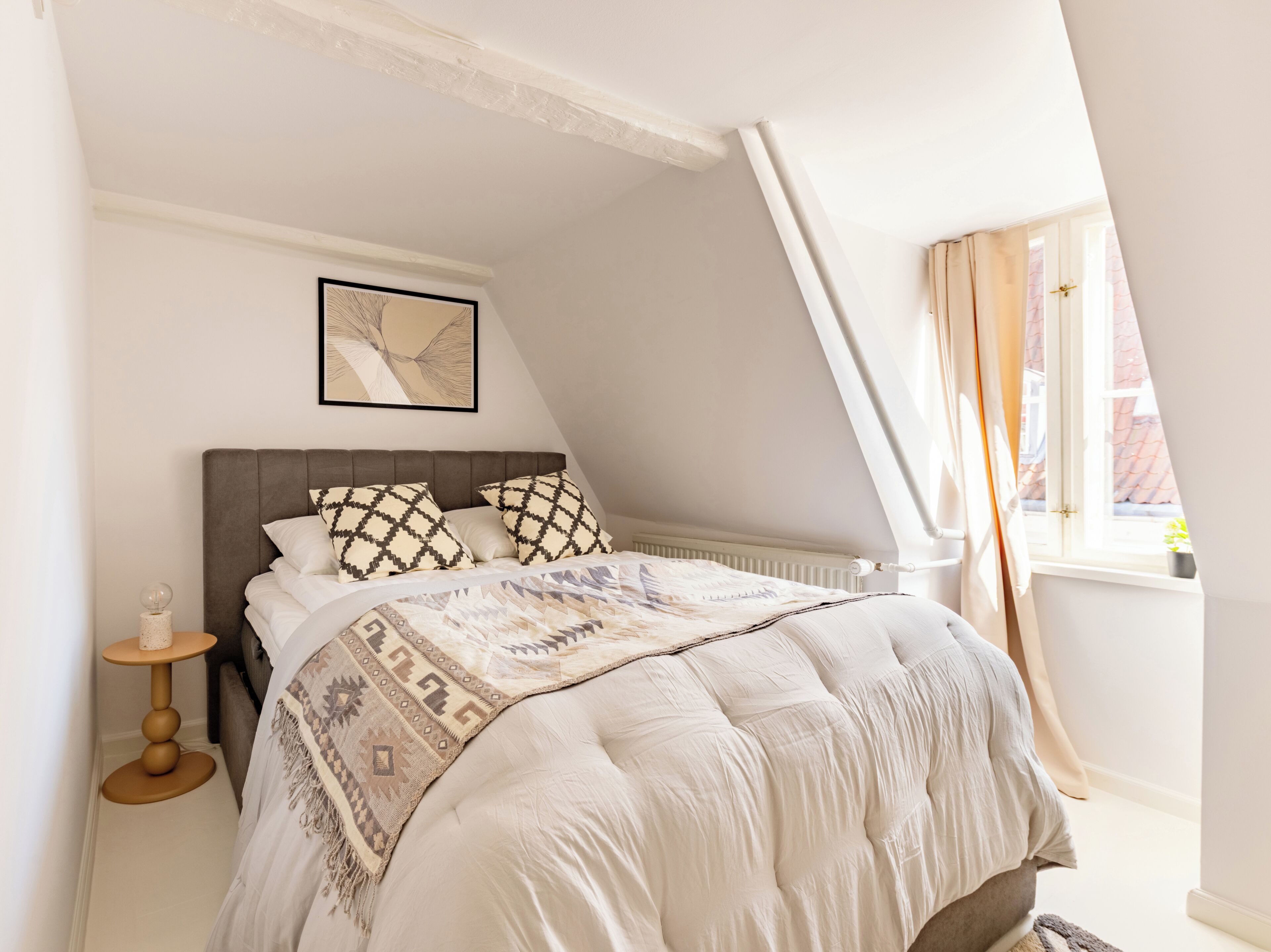 A bedroom with a sloped ceiling, a bed with patterned pillows, a bedside table with a lamp, and a window with curtains.