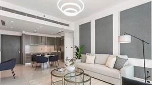 Un salon moderne avec un canapé, une table basse et des fauteuils. On aperçoit un coin cuisine au fond.