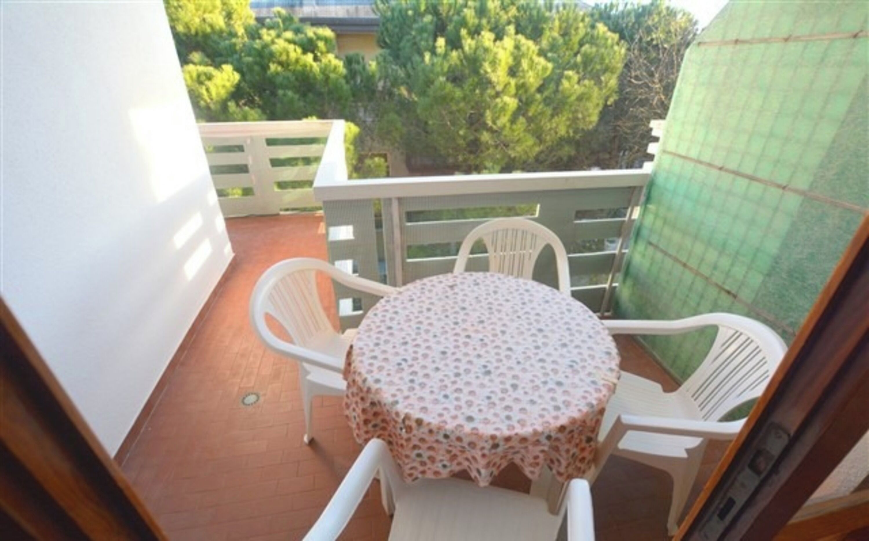 A small balcony with a round table and four white chairs, surrounded by a white railing and a green fence.
