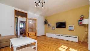 Uma sala de estar espaçosa com piso de madeira, um sofá branco, uma mesa de centro branca, uma televisão e estantes embutidas.