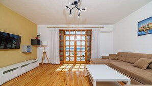 Uma sala de estar moderna com piso de madeira, mesa de centro branca, sofá bege, uma janela grande com cortinas e um quadro na parede.