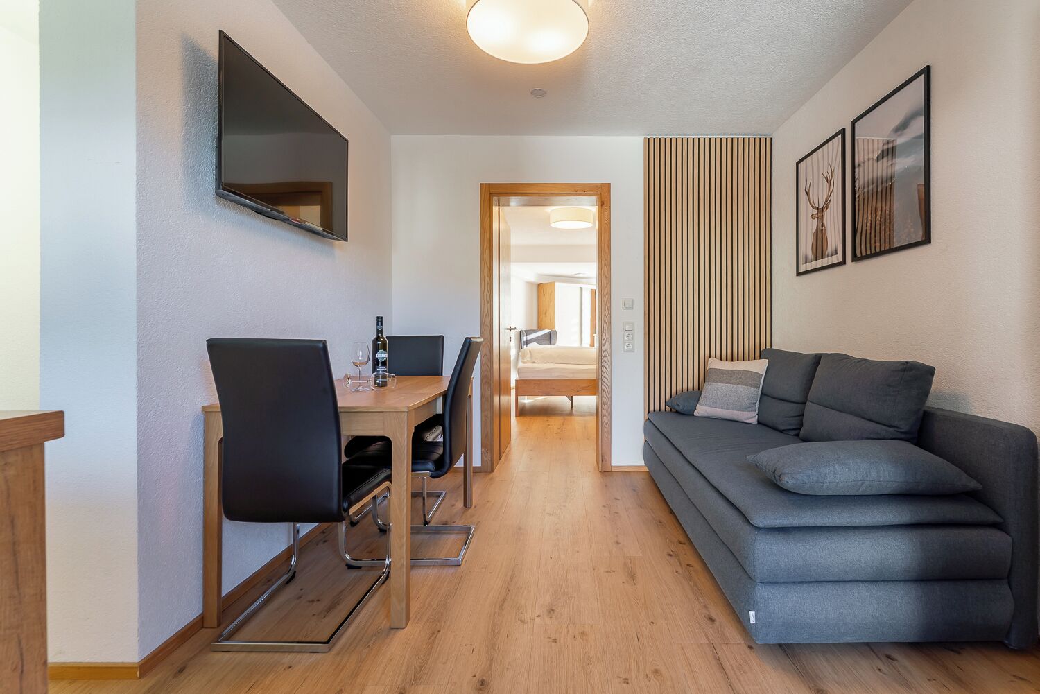 Un salon moderne avec un canapé gris, une table à manger en bois avec des chaises, un téléviseur fixé au mur et des tableaux encadrés accrochés au mur.