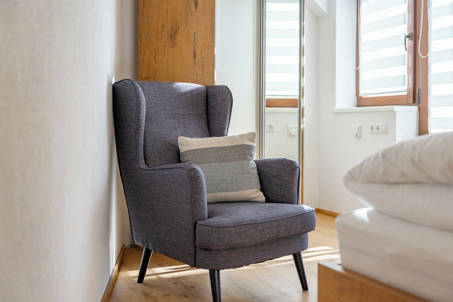A modern armchair with a cushion, positioned in a room with wooden flooring and a large window.