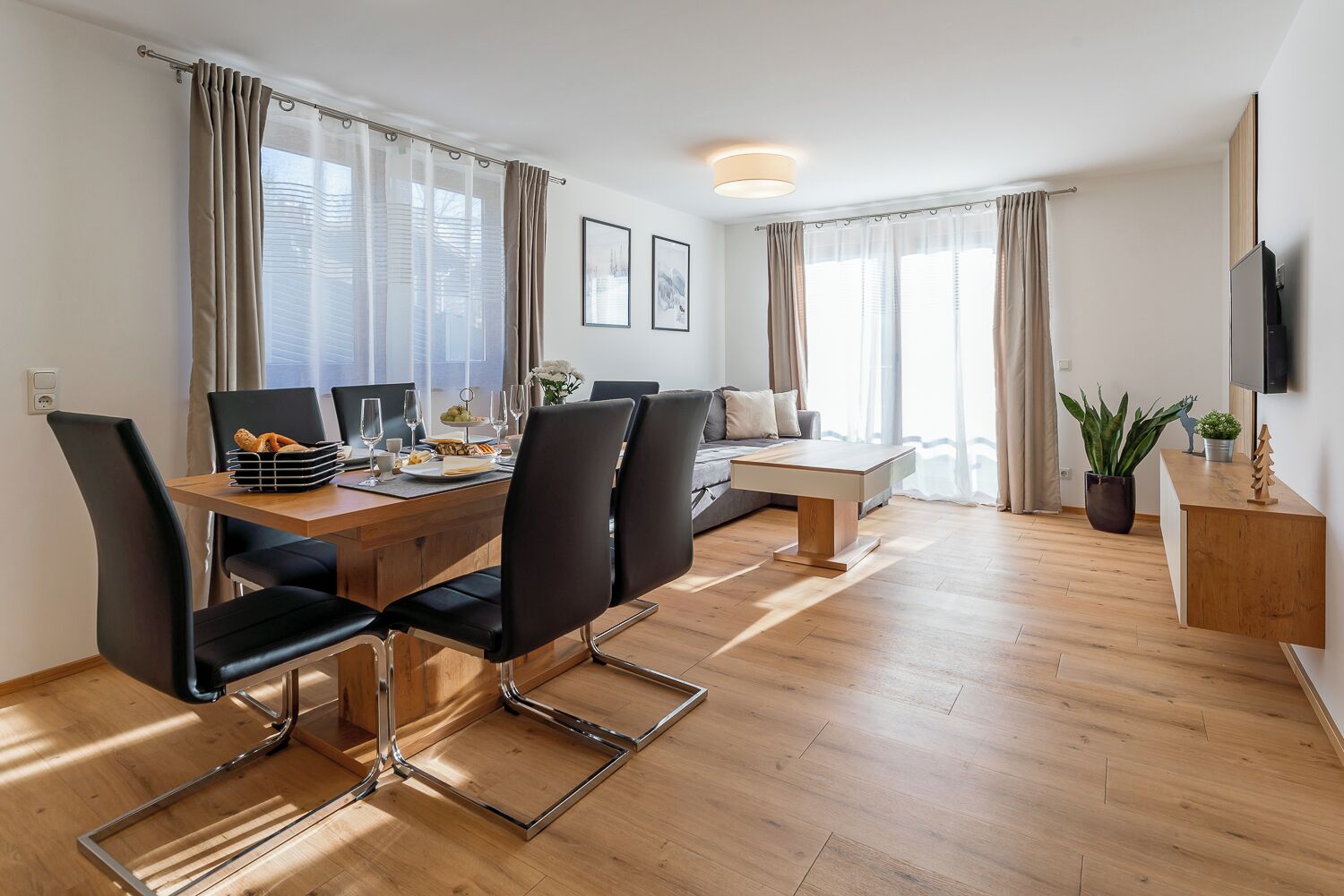 Uma sala de jantar moderna com uma mesa de madeira, cadeiras pretas e um sofá. Há quadros nas paredes, um vaso de planta e uma TV de tela plana.