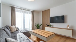 Ein modernes Wohnzimmer mit grauem Sofa, einem Holztisch, einem flachbildfernseher an einer Holzvertäfelung und einer kleinen Pflanze auf der Anrichte.